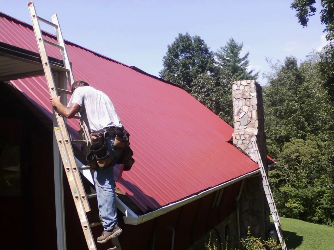 Roofer on a ladder beside a red metal roof and stone chimney — Soffit & Fascia Repair by Roofing Brothers in Coal Creek